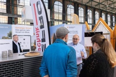 Drei Personen sprechen mit einem Mann an einem Messestand zum Thema Energie. Der Stand zeigt Banner und Informationsposter über intelligente und erschwingliche Elektrizität. Die Umgebung scheint ein Innenraum mit großen Fenstern im Hintergrund zu sein.