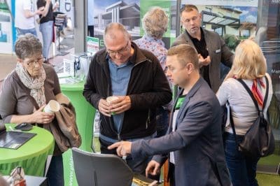 Eine Gruppe von Menschen steht bei einer Veranstaltung in einem Gebäude um einen grünen Tisch herum und unterhält sich. Ein Mann im Anzug zeigt auf einen Bildschirm, während andere zuhören und Tassen halten. Im Hintergrund sind Bilder von modernen Häusern zu sehen.