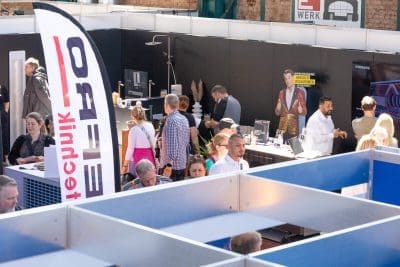 Auf einer belebten Technologiemesse in einer Halle gehen Menschen umher und tauschen sich aus. Stände mit elektronischen Anzeigen, Bannern und einem Pappausschnitt sind zu sehen. Die Atmosphäre ist lebhaft und professionell.