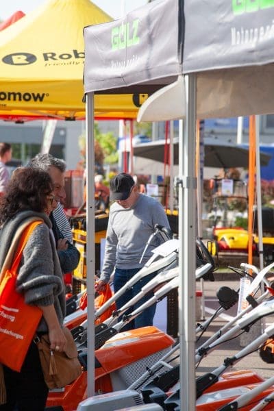 Menschen begutachten auf einer Messe oder Ausstellung Motorgeräte für den Außenbereich. Es sind orangefarbene und graue Maschinen ausgestellt, und im Hintergrund sind Markenzelte und Banner zu sehen. Die Szene wirkt geschäftig und lebendig.
