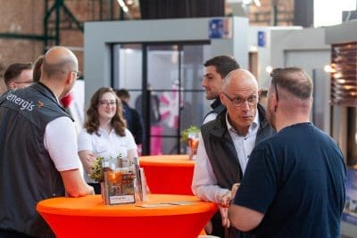 Bei einer Indoor-Veranstaltung stehen Menschen um orangefarbene Tische herum und unterhalten sich. Einige tragen Markenwesten oder weiße Hemden. Auf den Tischen liegen Broschüren und kleine Pflanzen, und im Hintergrund sind moderne Messestände oder Displays zu sehen.
