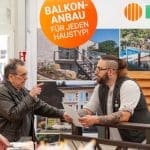 Ein älterer Mann unterhält sich mit einem bärtigen Mitarbeiter, der an einem Stand mit einem Schild "BALKON-ANBAU FÜR JEDEN HAUSTYP!" und Fotos von Balkonen im Hintergrund Papiere hält.