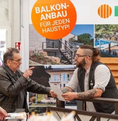 Ein älterer Mann unterhält sich mit einem bärtigen Mitarbeiter, der an einem Stand mit einem Schild "BALKON-ANBAU FÜR JEDEN HAUSTYP!" und Fotos von Balkonen im Hintergrund Papiere hält.