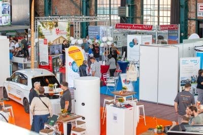 Eine geschäftige Messe in einer Halle mit Besuchern an verschiedenen Ständen, Informationsdisplays und einem ausgestellten weißen Auto. Helle Banner, Tische und Informationsplakate sind überall im Veranstaltungsraum zu sehen.
