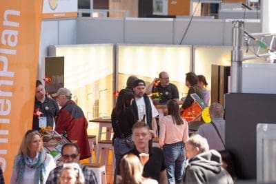 Eine Gruppe von Menschen, sowohl stehend als auch sitzend, interagiert bei einer Indoor-Veranstaltung oder Expo mit heller Beleuchtung und Informationsständen. Auf der linken Seite des Bildes ist ein orangefarbenes Banner zu sehen.