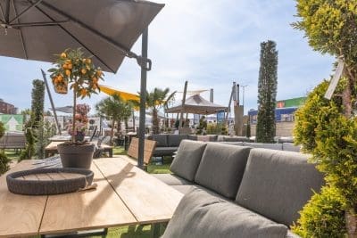 Outdoor-Möbelausstellung mit grauen Sofas, Holztischen, Topfpflanzen und einem Orangenbaum unter großen Sonnenschirmen an einem sonnigen Tag in einem Gartencenter oder Outdoor-Ausstellungsraum.