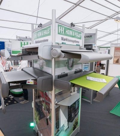 Ein Messestand mit verschiedenen versenkbaren Markisensystemen von HEIM & HAUS, mit ausgefahrenen Mustermarkisen in verschiedenen Farben und Stilen, unter einem weißen Zelt auf einer Ausstellung.