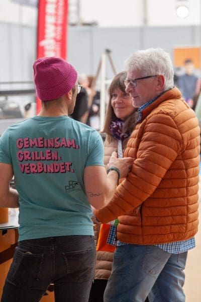Ein Mann mit rosa Mütze und türkisfarbenem Hemd mit deutscher Aufschrift unterhält sich mit einem älteren Mann in einer orangefarbenen Jacke und einer Frau in einem Haus. Auf dem Hemd steht GEMEINSAM. GRILLEN. VERBINDET. (Zusammen. Grillen. Verbinden.).