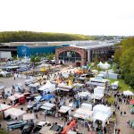 Luftaufnahme einer Freiluftmesse mit weißen Zelten, Fahrzeugen und Menschen, die sich vor einem großen Industriegebäude aus Backstein versammelt haben, das von Grünflächen umgeben ist.
